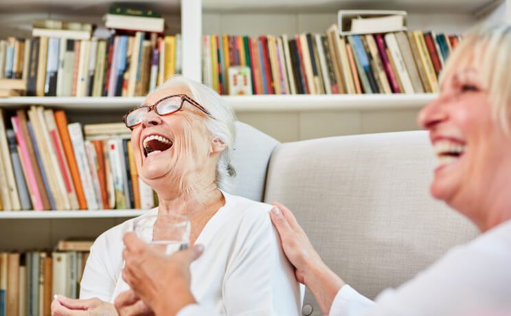 Two elderly women are laughing together in front of a bookshelf.