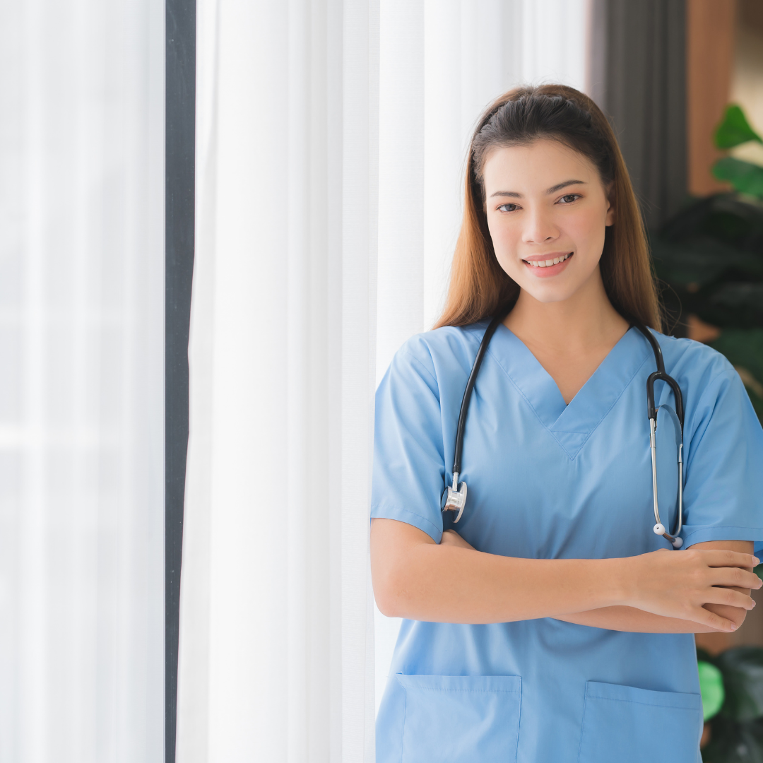 A nurse with a stethoscope around her neck is standing in front of a window with her arms crossed.