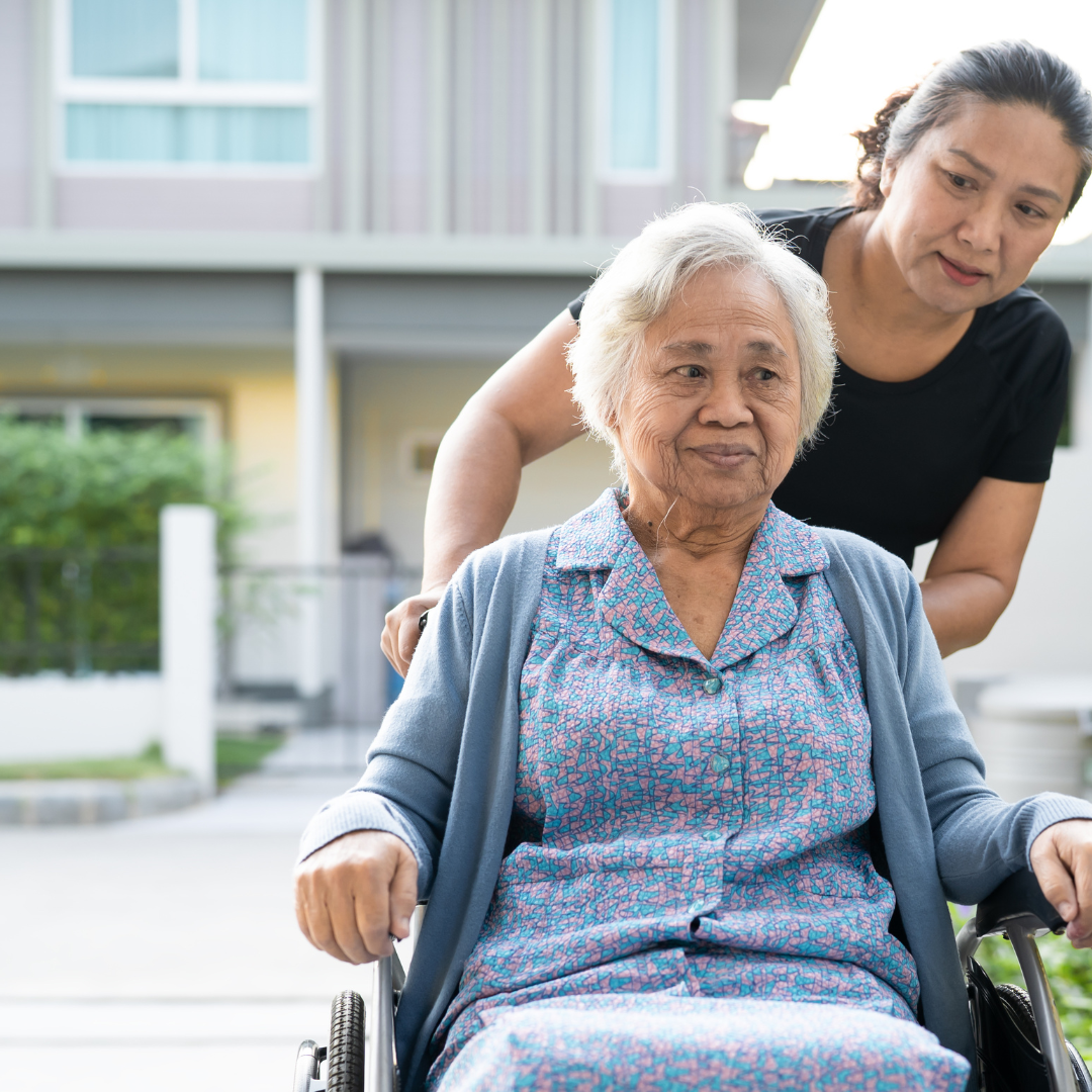 a caregiver pushes an elderly woman outside in a wheelchair on a nice day.