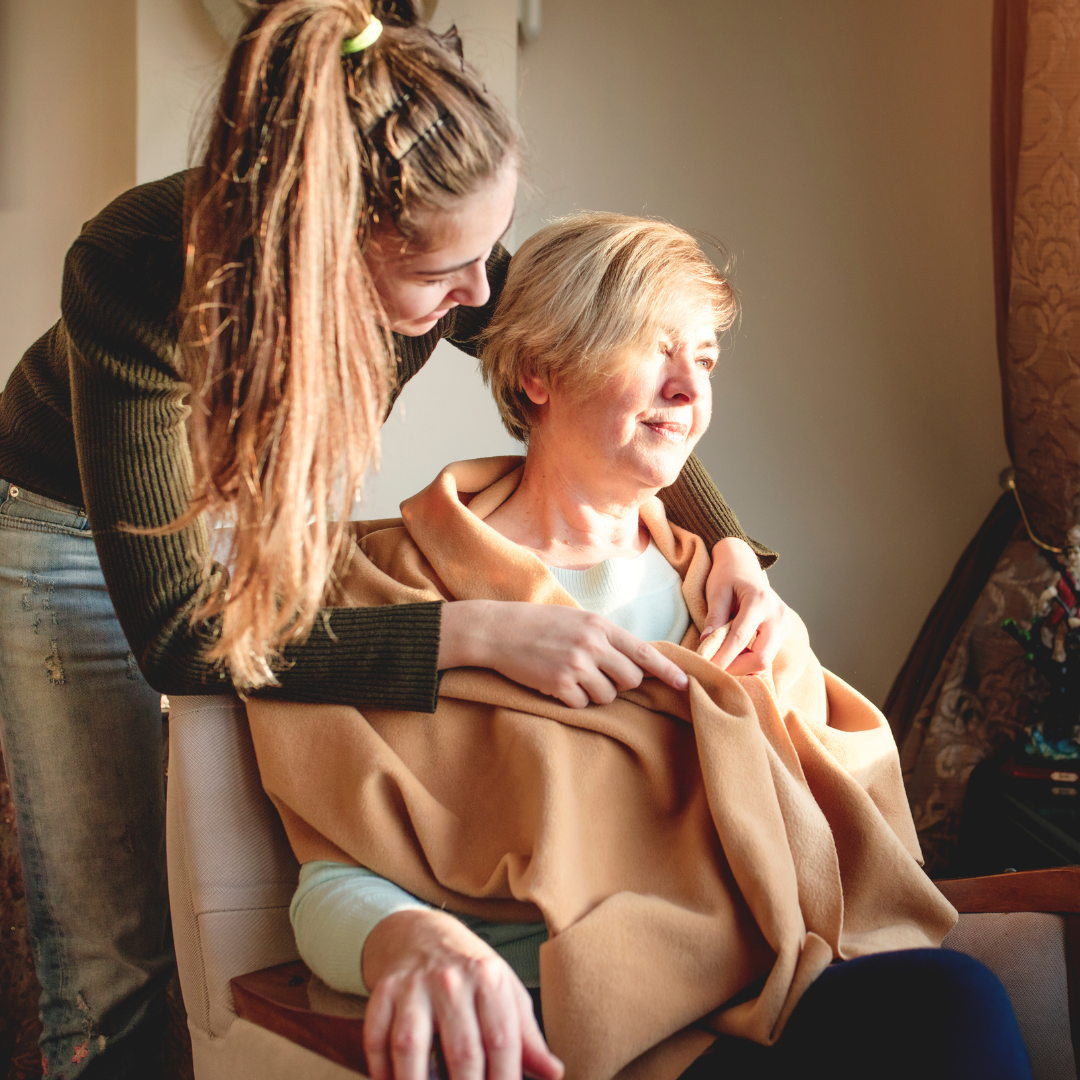 A woman is putting a blanket on an older woman who is sitting in a chair