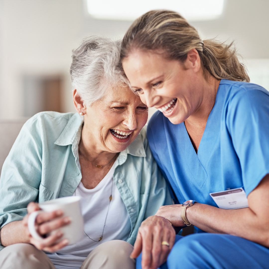 A nurse and an older woman are laughing together while sitting on a couch.