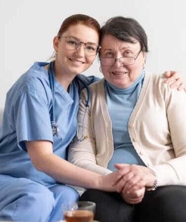 A nurse and an elderly woman are sitting on a couch holding hands.