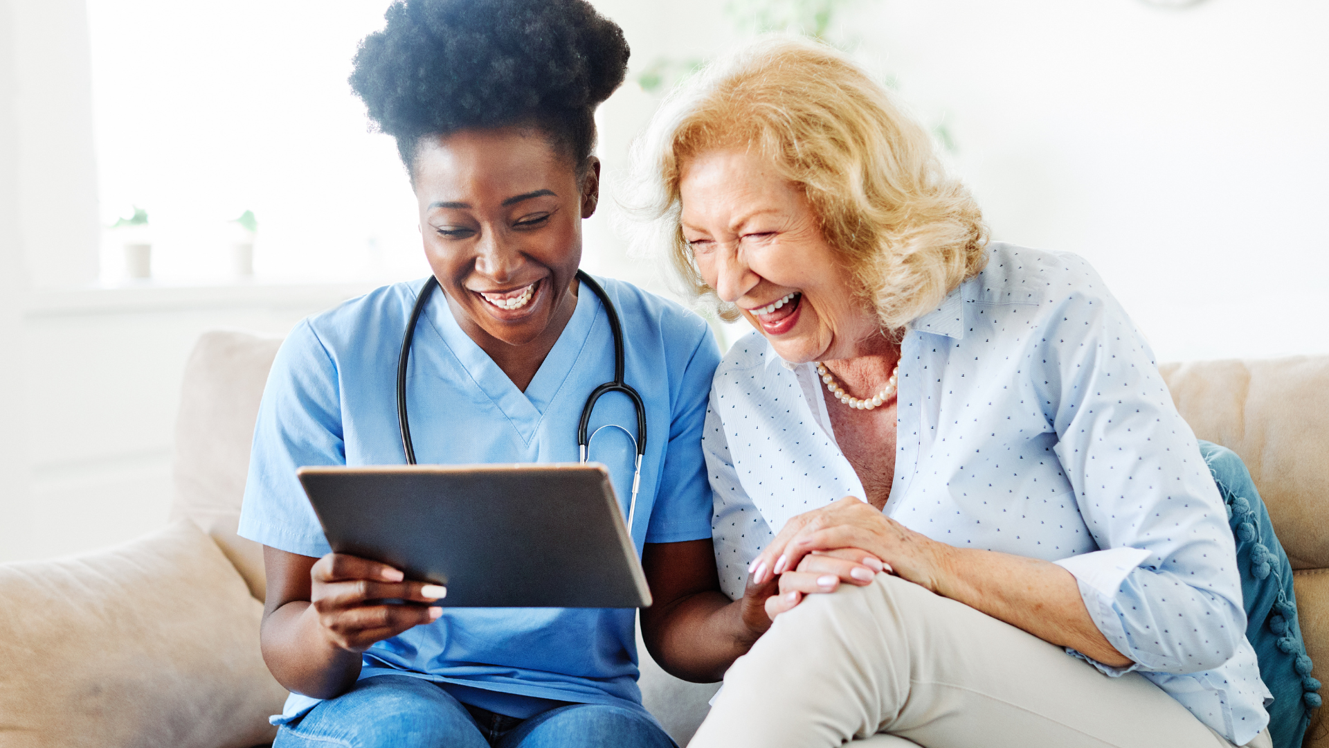 A caregiver laughs with an Alzheimers patient while watching a video on a tablet.