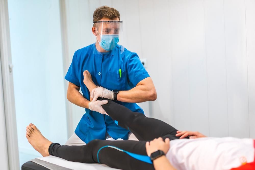 A Man Wearing a Mask and Gloves Is Stretching a Patient's Leg — Action Physiotherapy in Cooks Hill, NSW