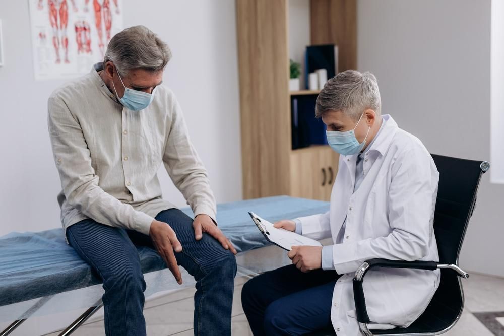 A Doctor Is Examining a Patient's Knee While Wearing a Mask — Action Physiotherapy in Cooks Hill, NSW