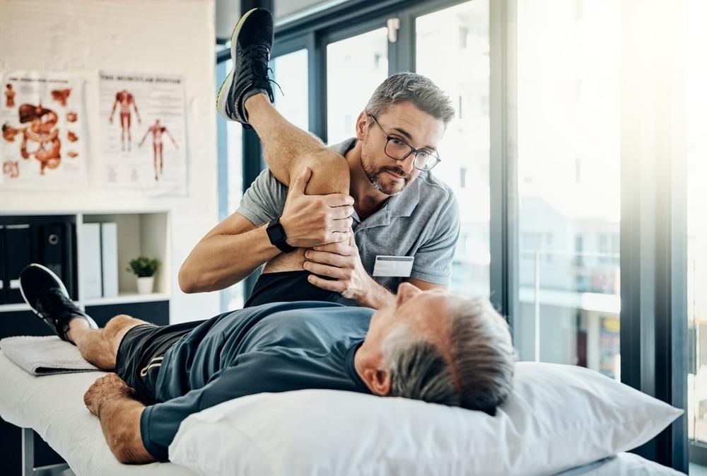 A Man Is Laying on A Bed While a Physical Therapist Stretches His Leg — Action Physiotherapy in Cooks Hill, NSW