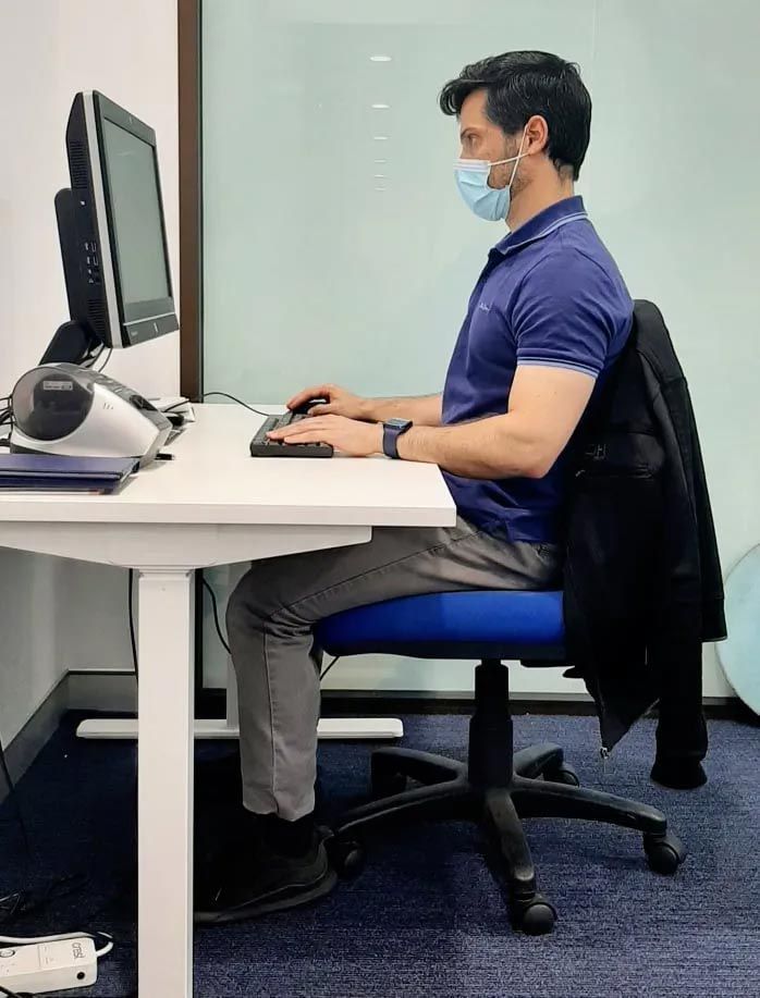 A Man Wearing a Mask Is Sitting at A Desk in Front of A Computer — Action Physiotherapy in Cooks Hill, NSW