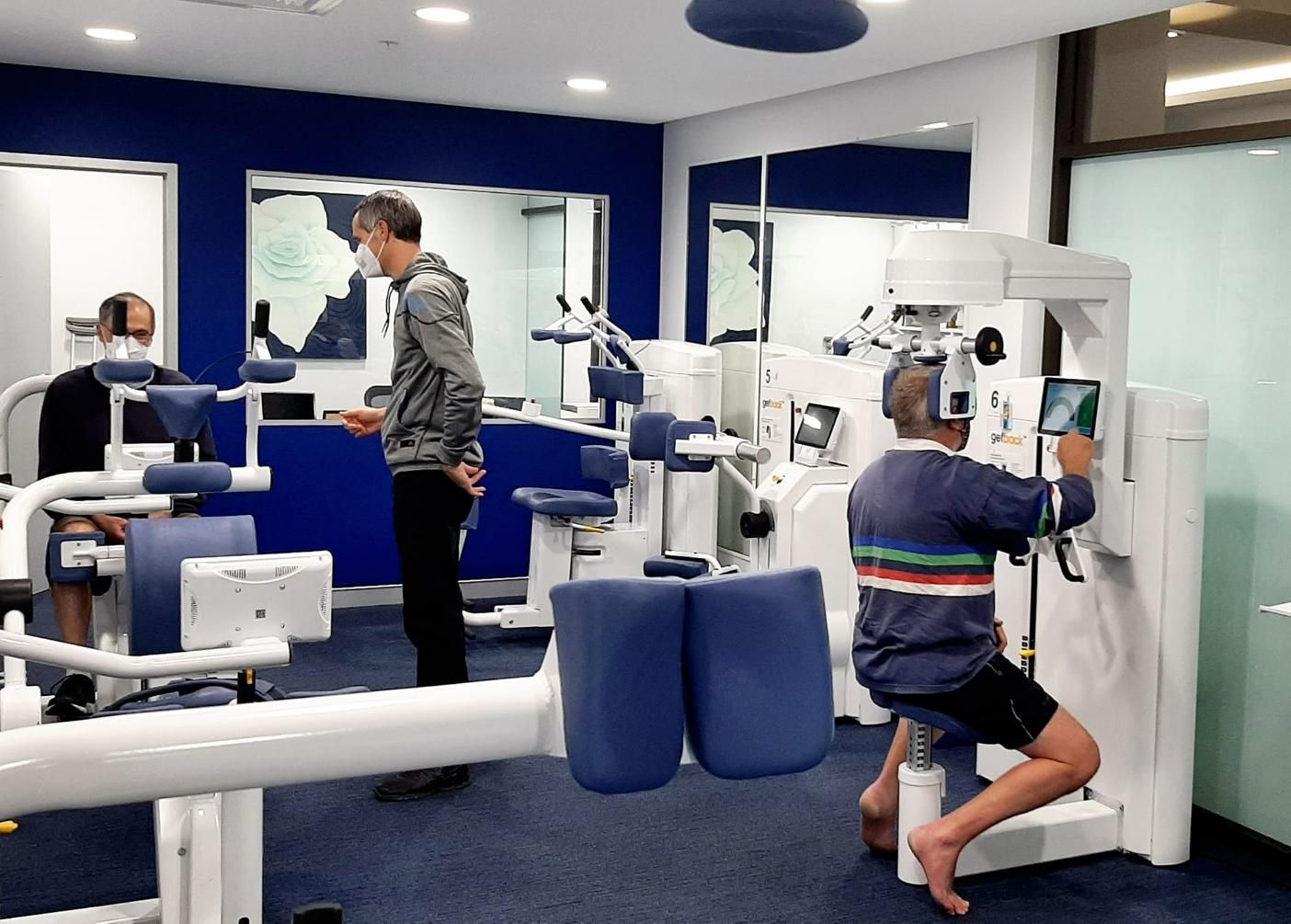 A Man Is Using a Machine in A Gym While Another Man Looks On — Action Physiotherapy in Cooks Hill, NSW