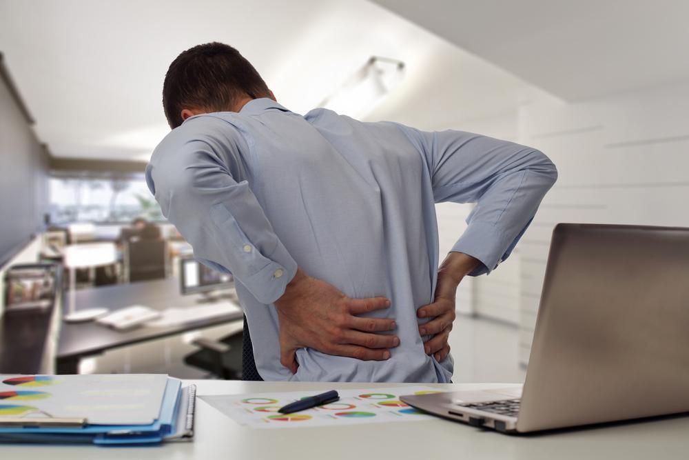 A Man Is Sitting at A Desk with His Hands on His Back — Action Physiotherapy in Cooks Hill, NSW