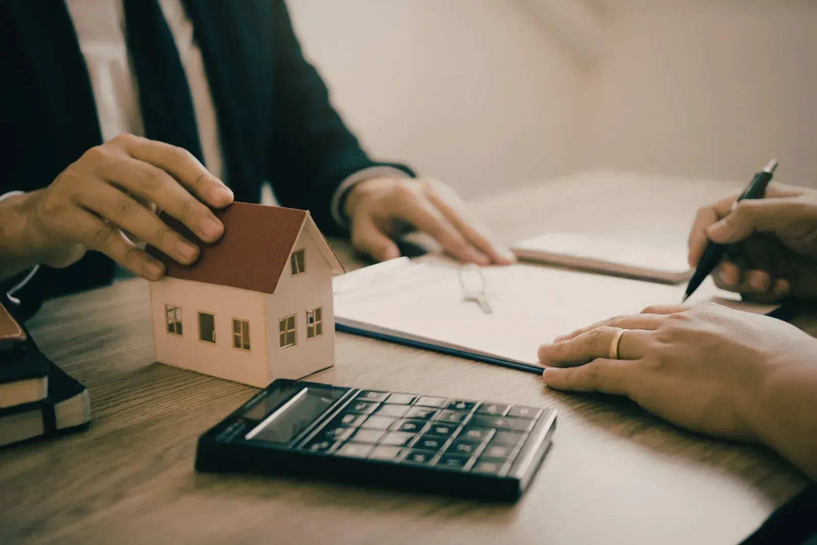 Hands in a suit and business attire touch a model house while signing a contract at a desk with a calculator nearby.