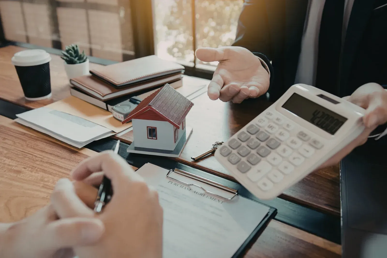 A professional meeting at a wooden desk with a model house, a calculator, documents, and a pen.
