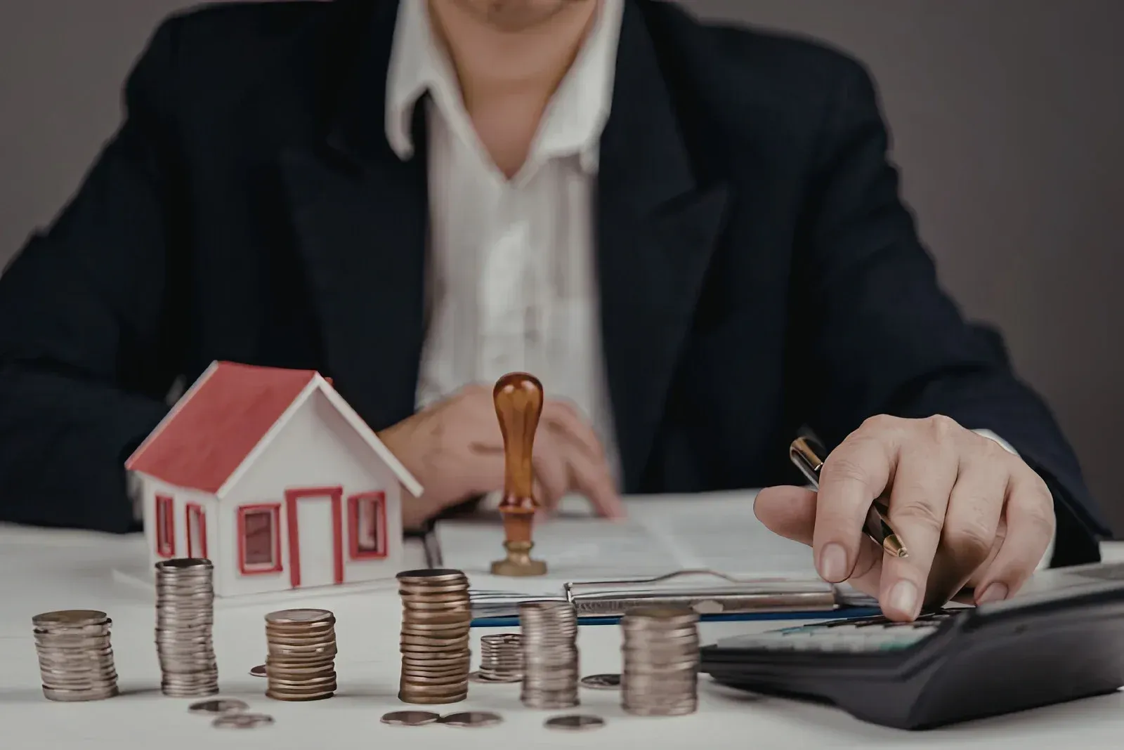 A person in a business suit calculates finances next to a model house, a wax seal, and stacks of coins.