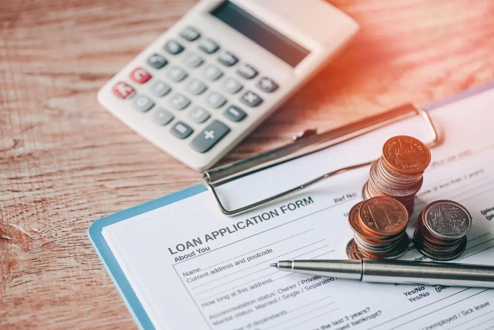 A loan application form on a clipboard with a pen, a stack of coins, and a calculator on a wooden table.