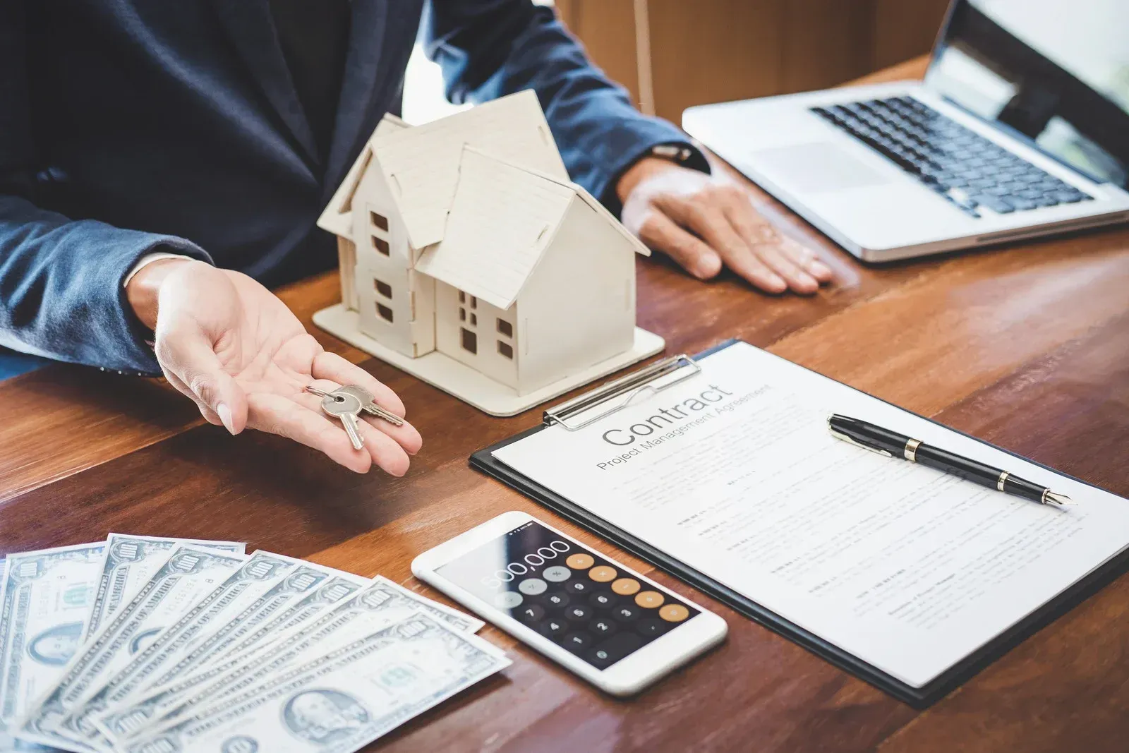 A person holds house keys on a desk with a model house, a contract, a calculator, and a stack of cash.