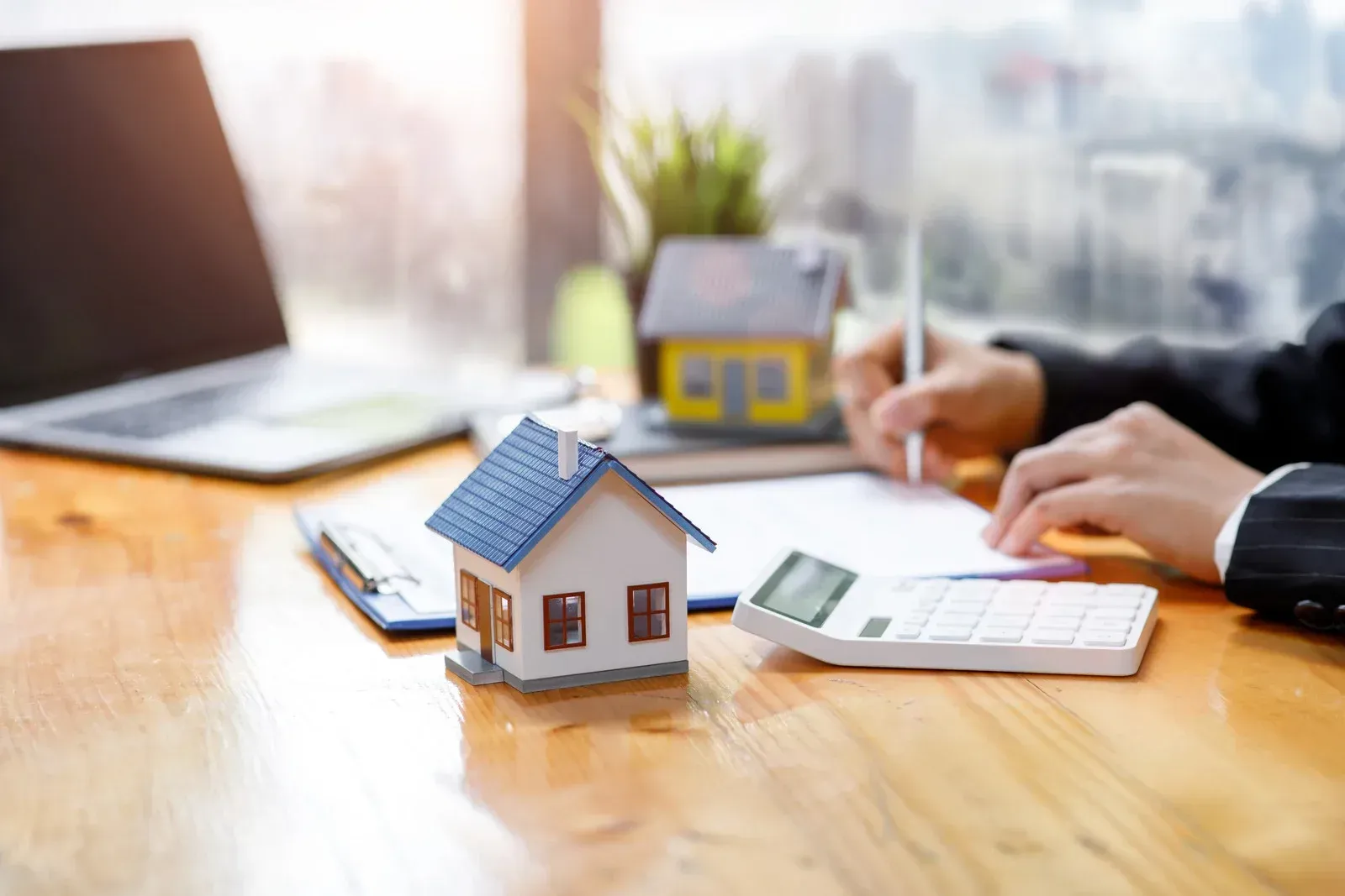 A person writing at a desk with a laptop, a calculator, and two model houses, representing a real estate transaction.