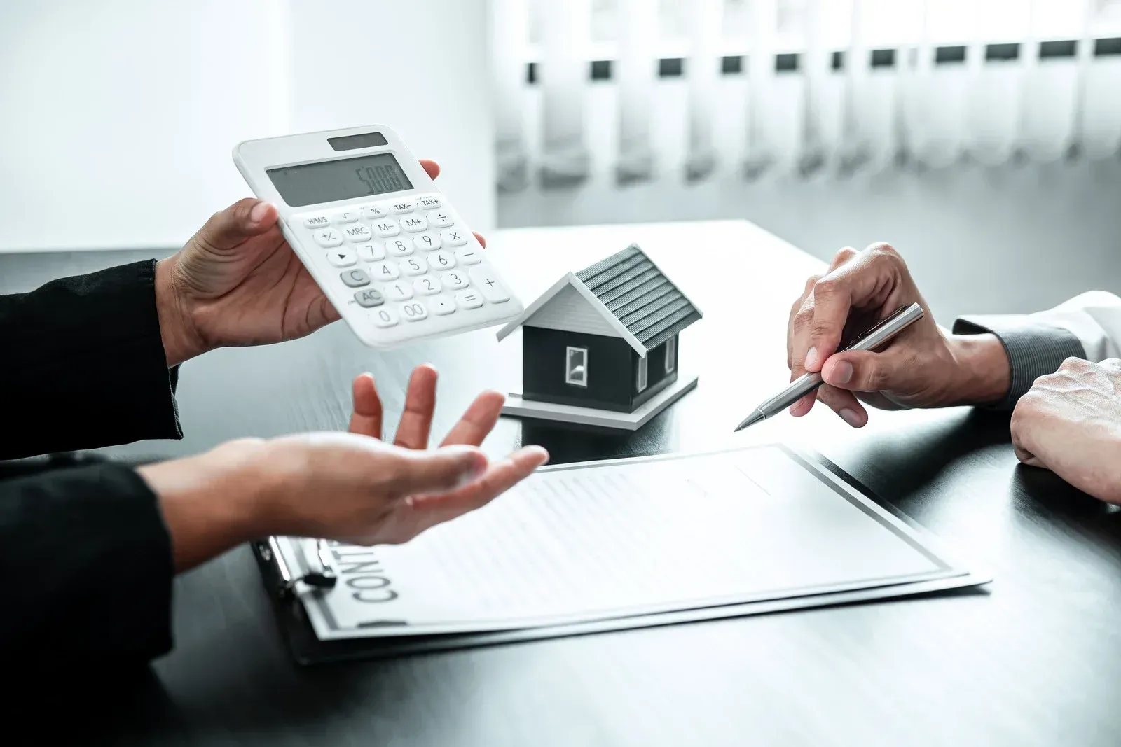 Two people at a desk reviewing a house model and a contract with a calculator, discussing real estate or insurance.