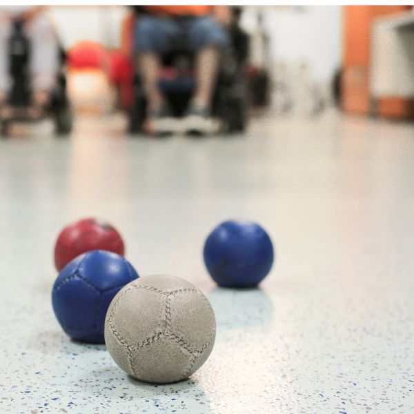 four boccia balls one red, two blue and one white sit close to one another in the foreground. two people using wheelchairs are in the background 