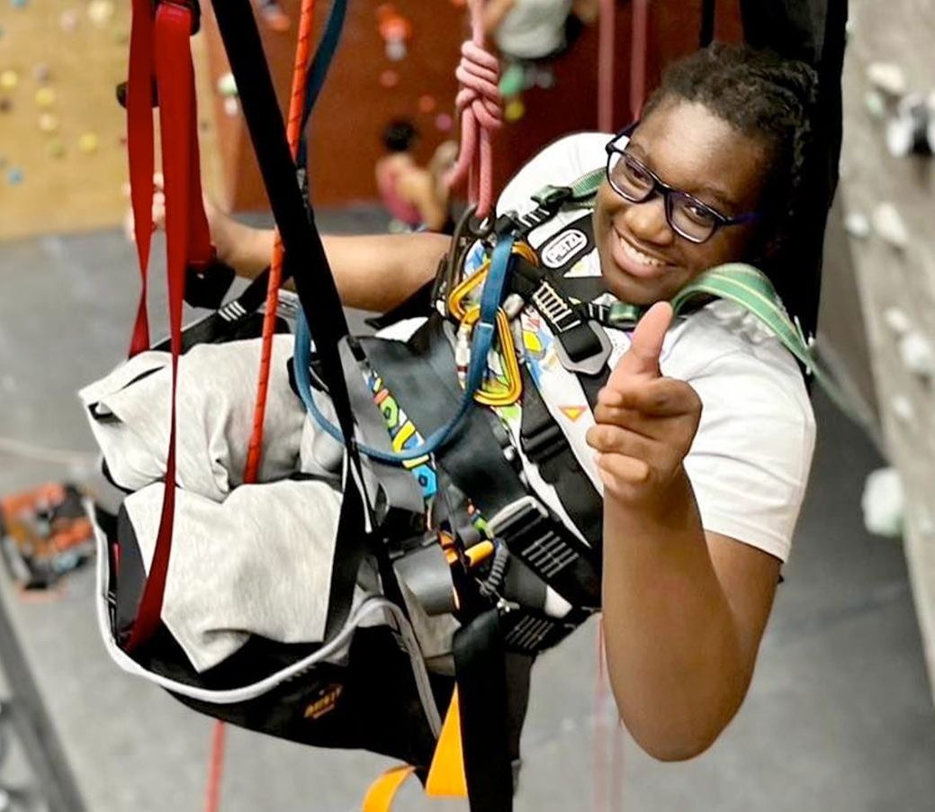 Climber with double leg amputation smiling and pointing while hanging from an indoor rock wall with safety ropes