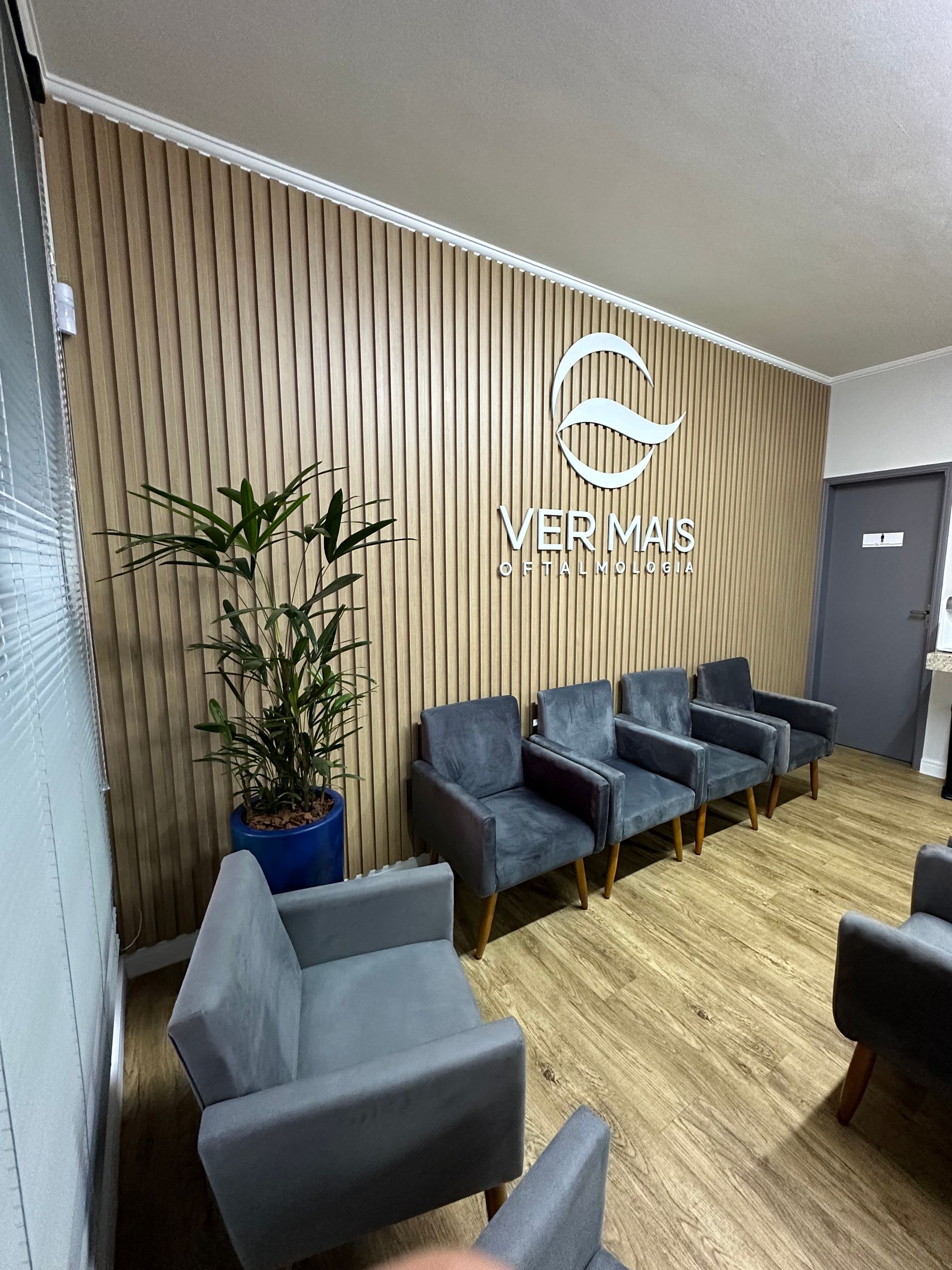 Sala de espera com poltronas cinza, planta e parede com painel de madeira e logotipo Ver Mais