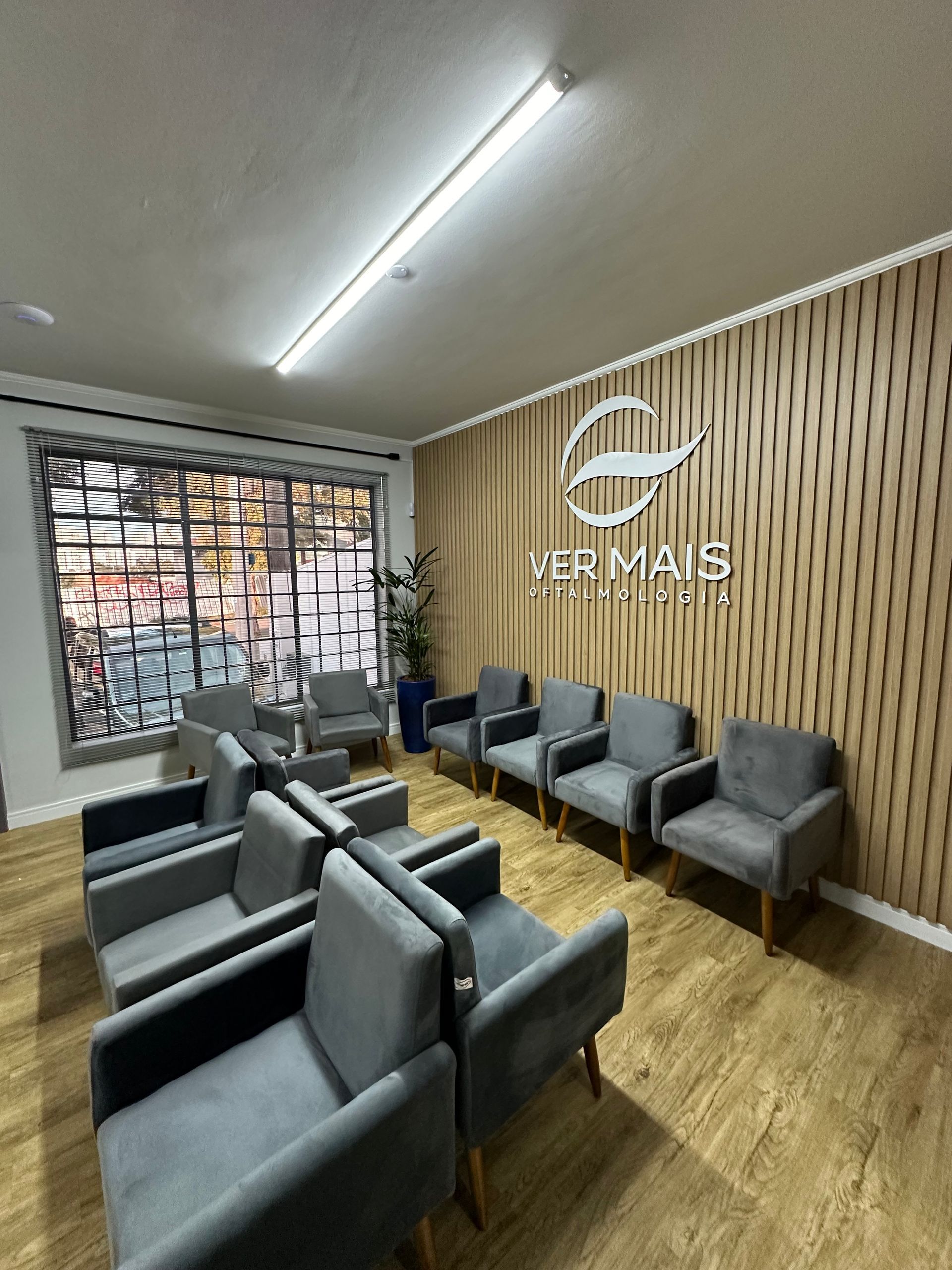 Sala de espera com cadeiras cinza, piso imitando madeira e parede texturizada com o logotipo.