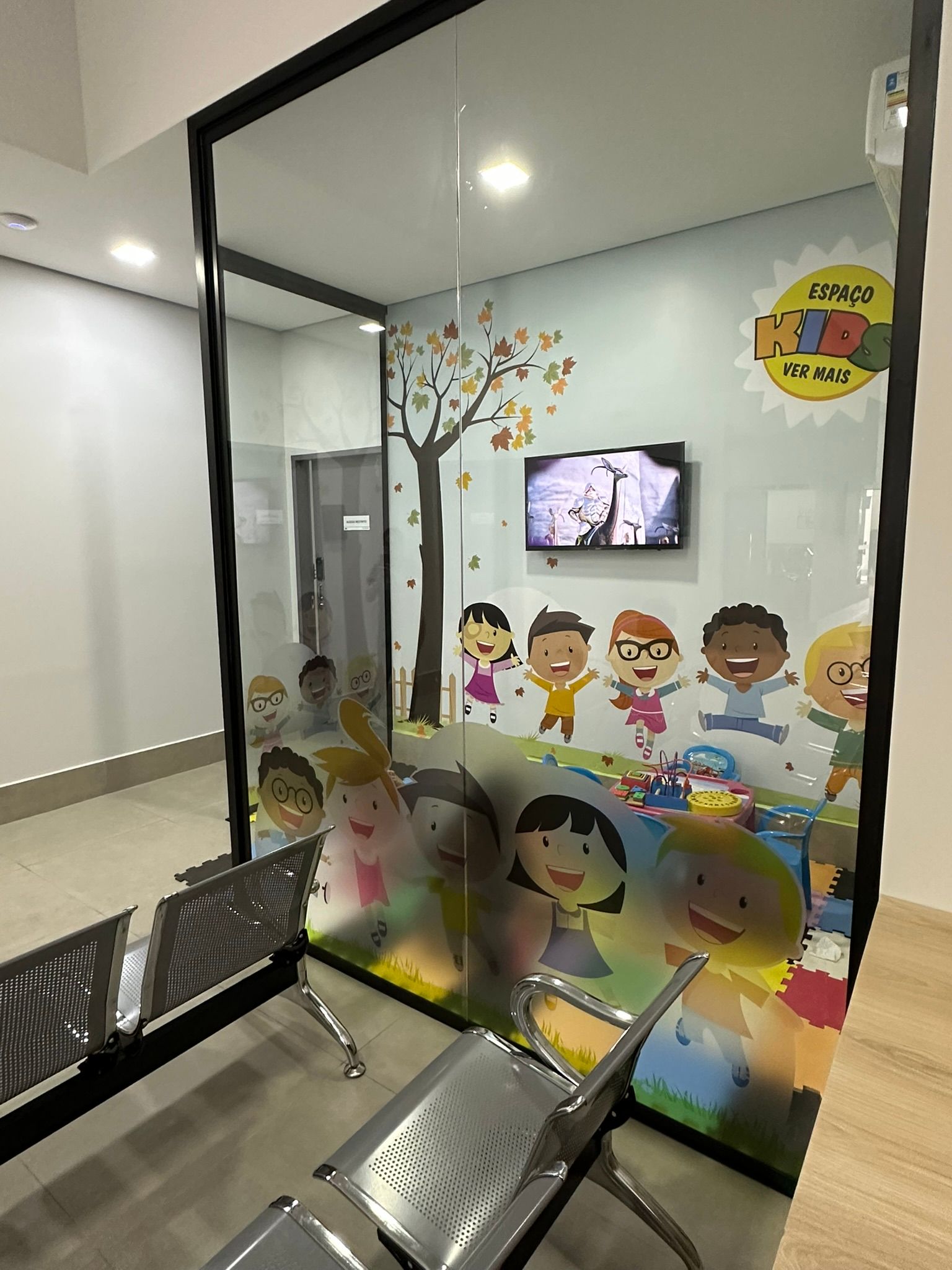 Sala de espera com parede de vidro decorada com desenhos animados infantis, área de estar visível.