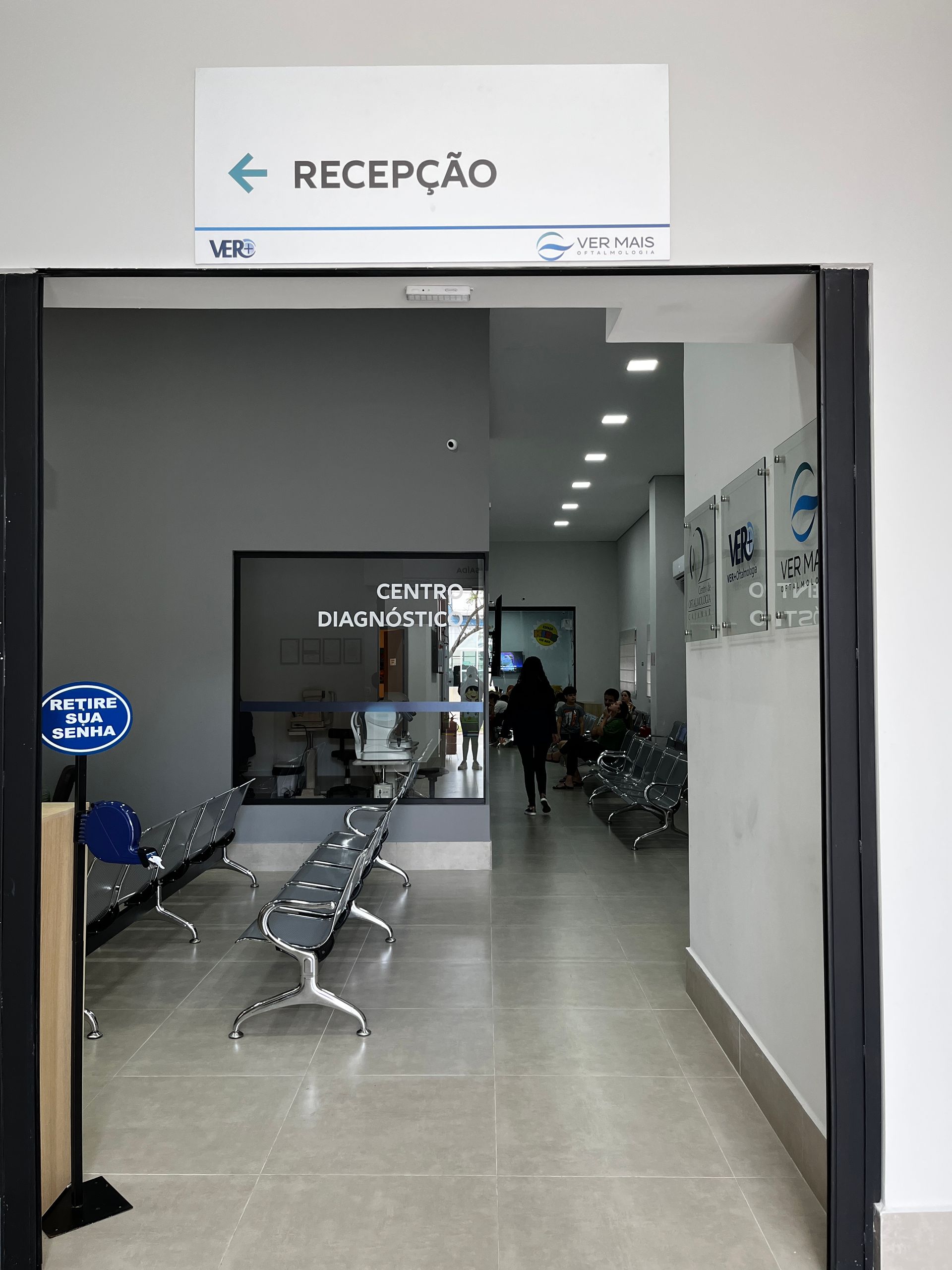Corredor que leva à recepção, com cadeiras de espera. Placa: Recepção. Pessoa se afastando.