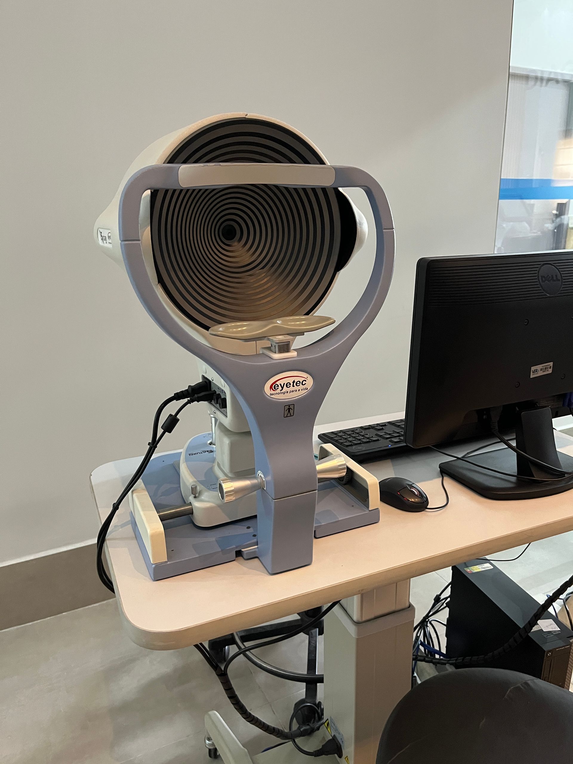 Aparelho para exame oftalmológico com alvo de círculos concêntricos, mouse e computador sobre uma pequena mesa.