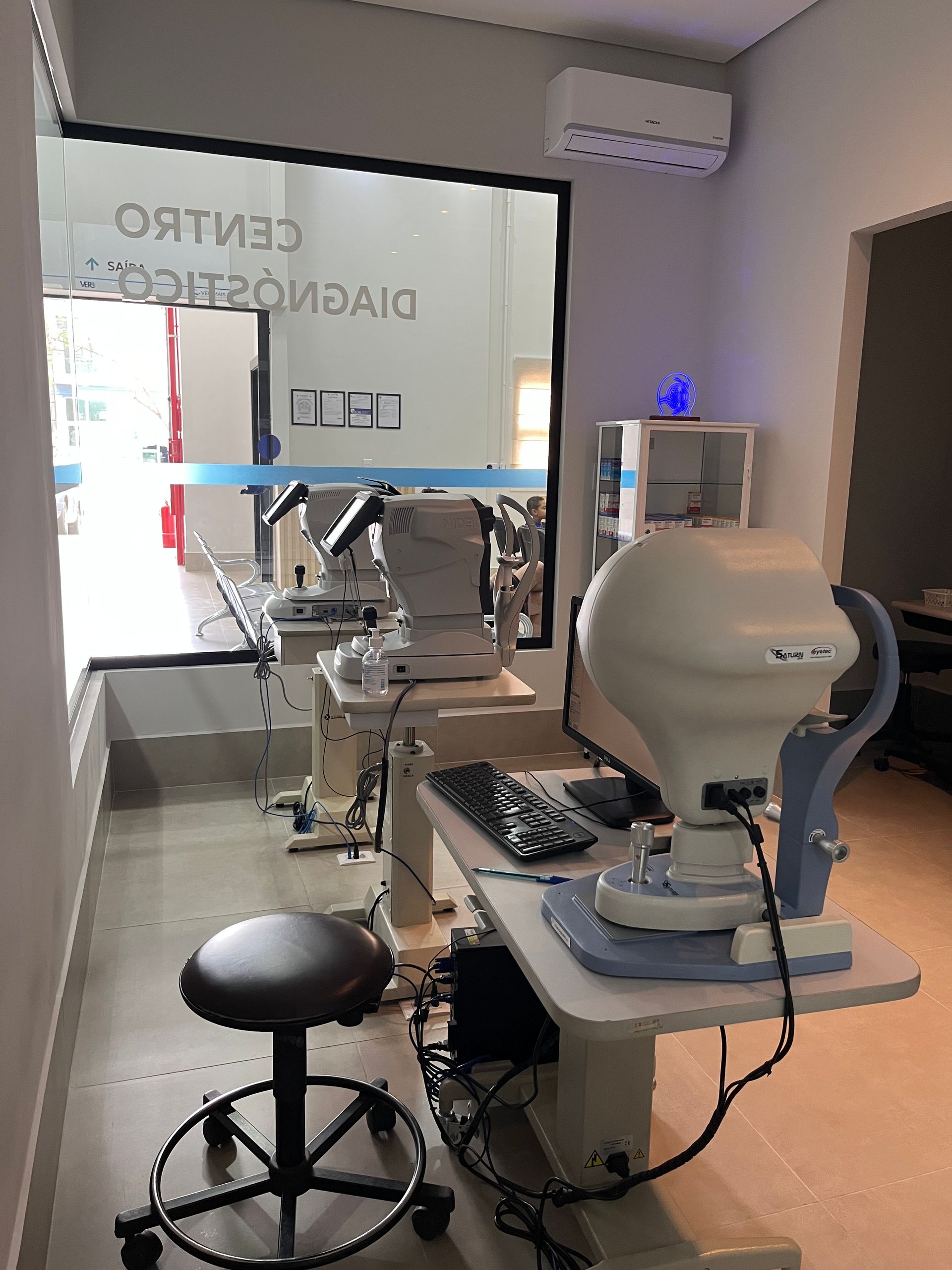 Equipamentos de optometria em uma sala de clínica, incluindo instrumentos, um banco e um computador.