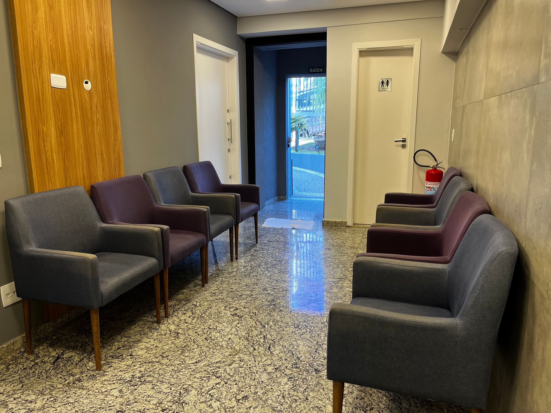 Sala de espera com poltronas em tons de cinza e roxo, de frente para um corredor.