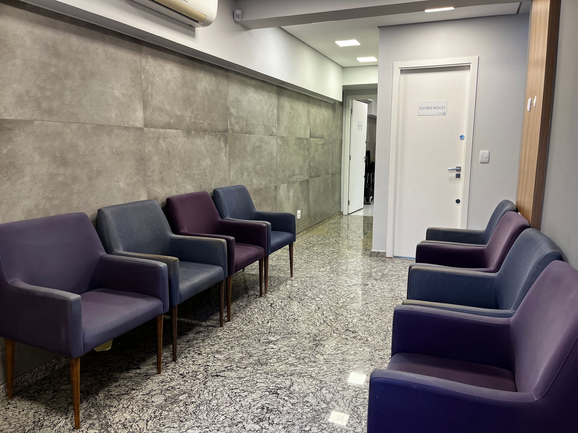 Sala de espera com fileiras de cadeiras roxas e azuis encostadas em uma parede de pedra, com uma porta branca ao fundo.