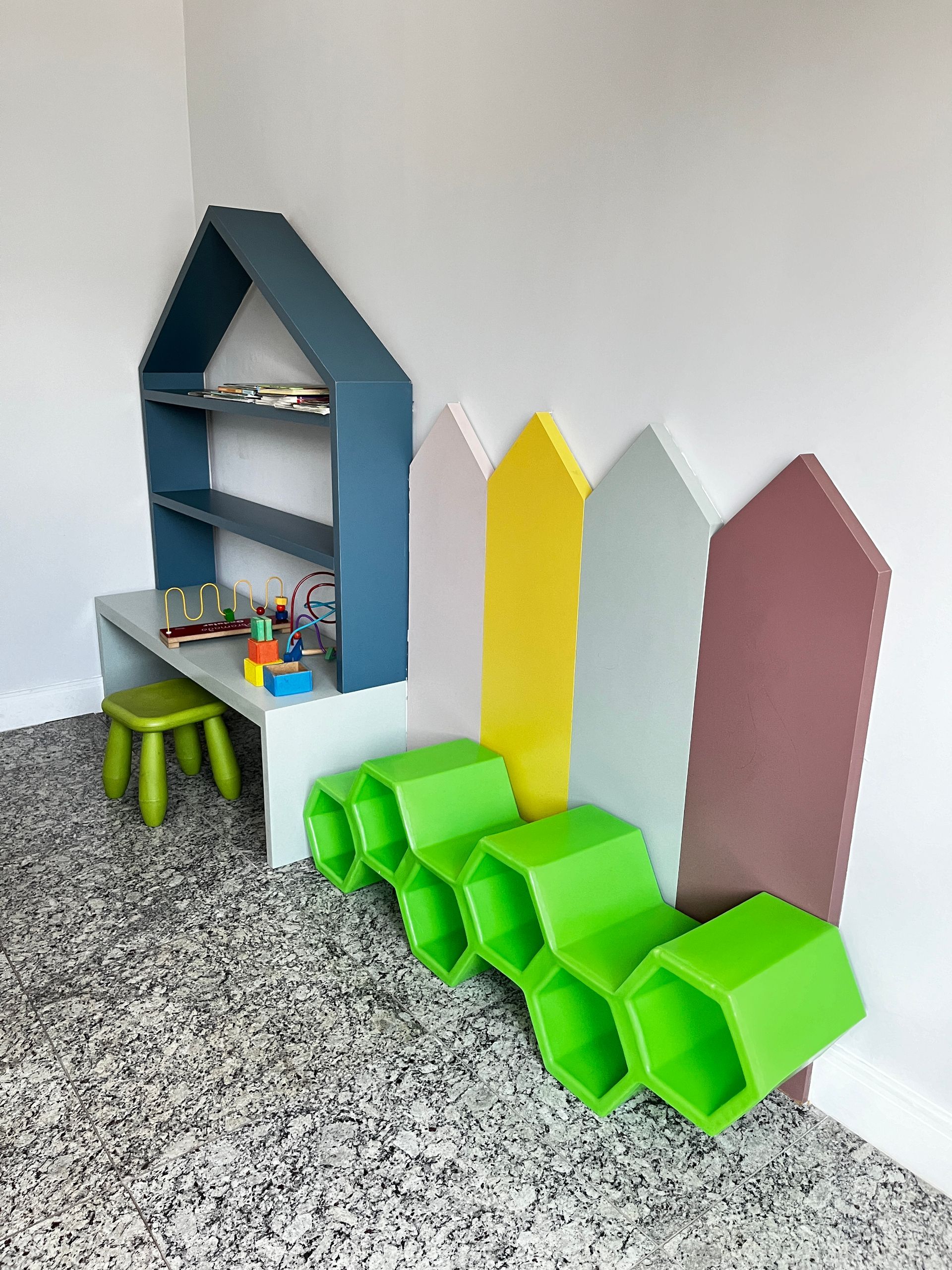 Área de recreação: prateleira azul em formato de casa, banquinho verde, , cerca colorida e brinquedos.