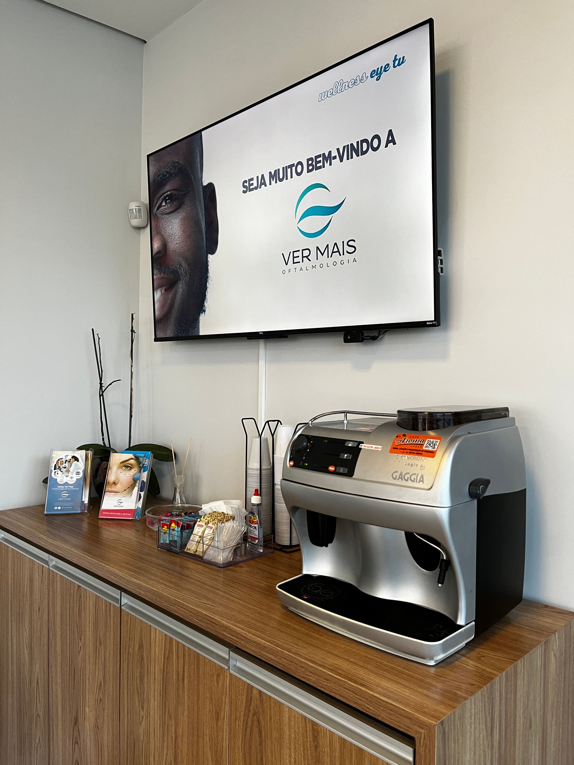 Máquina de café e balcão na área de espera com publicidade na TV.