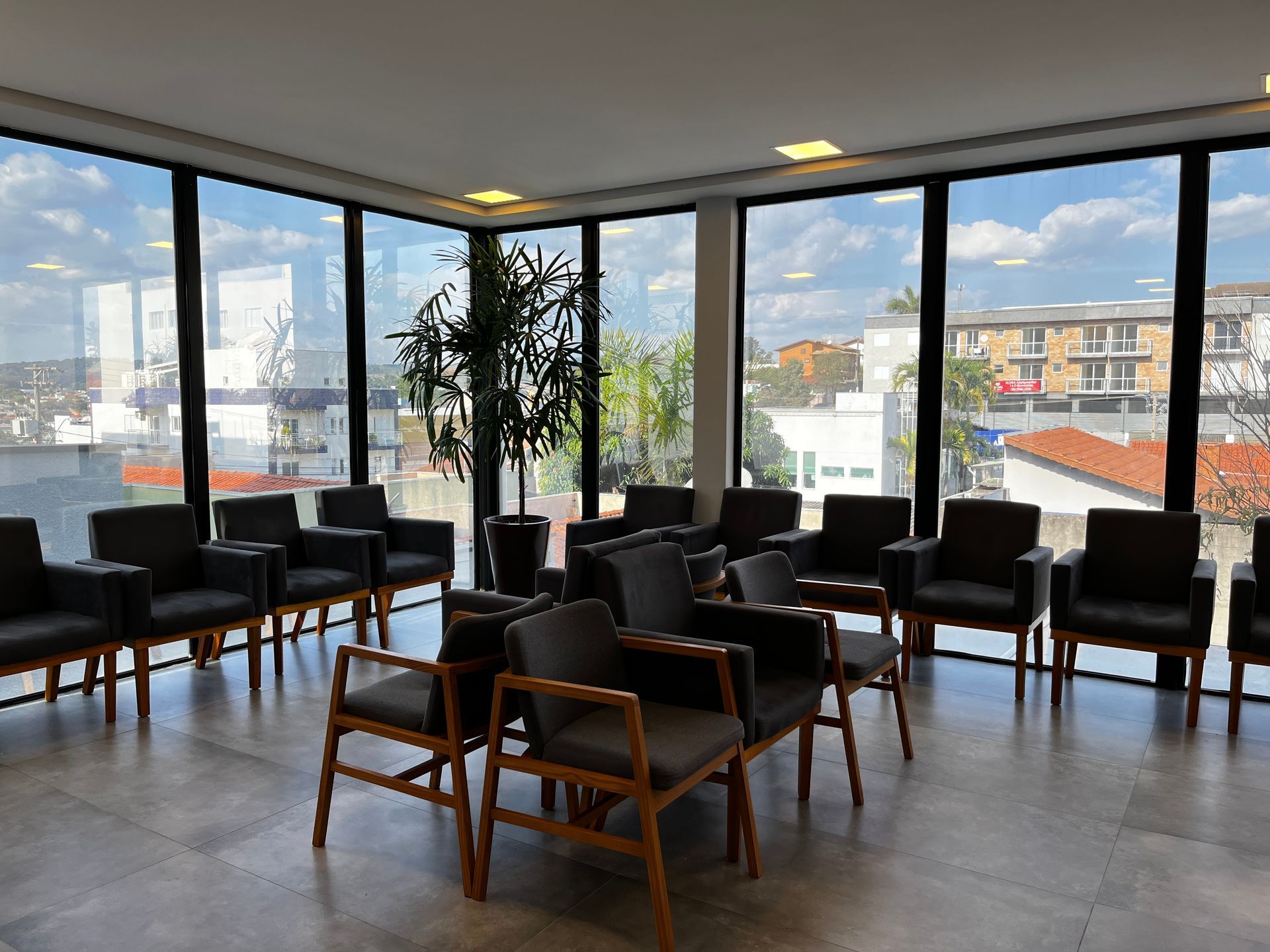 Sala de espera com muitas cadeiras escuras, janelas grandes e uma árvore de interior.