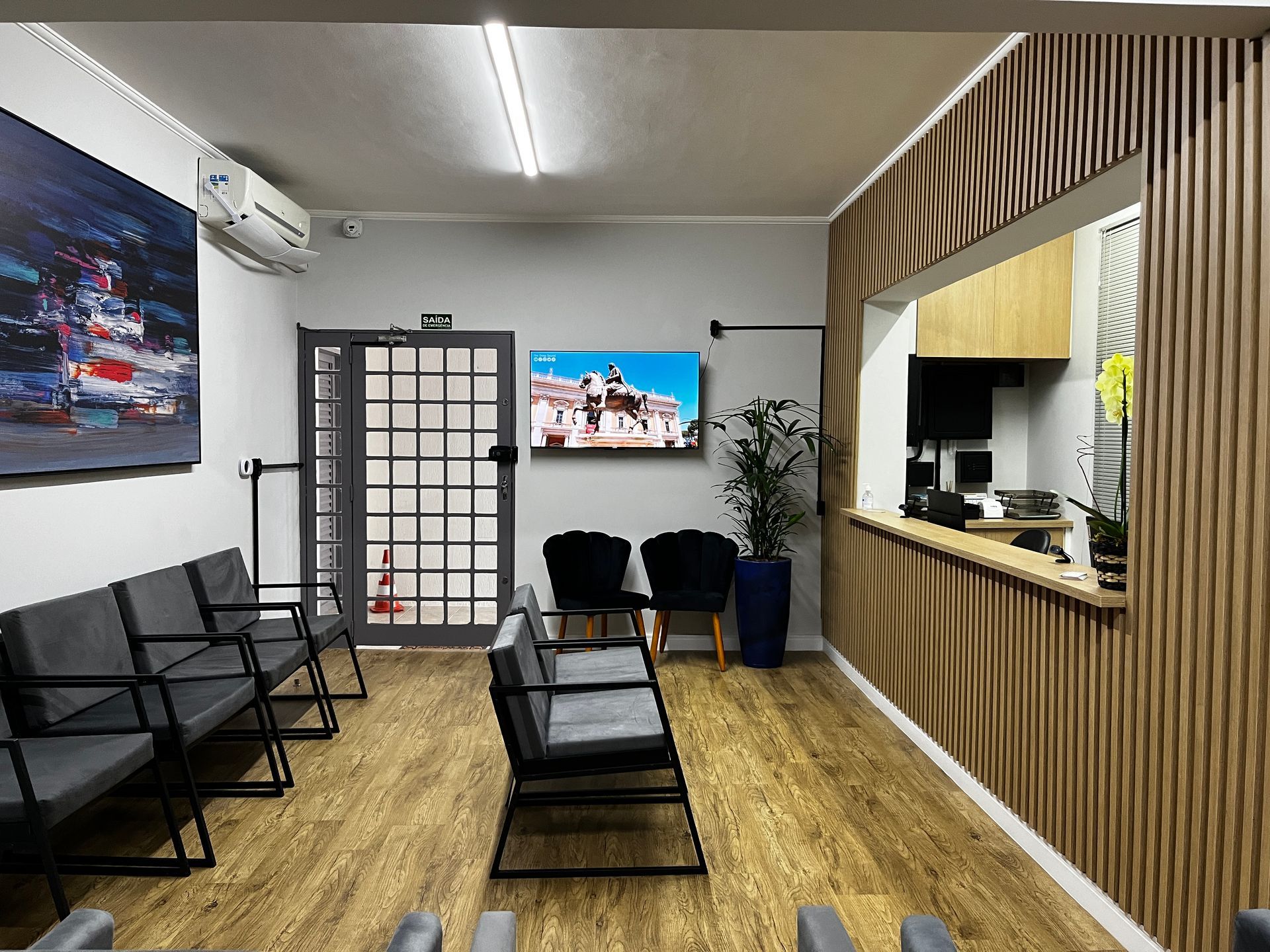 Sala de espera com assentos, obras de arte e área de recepção. Piso e paredes de madeira; paleta de cores neutras.