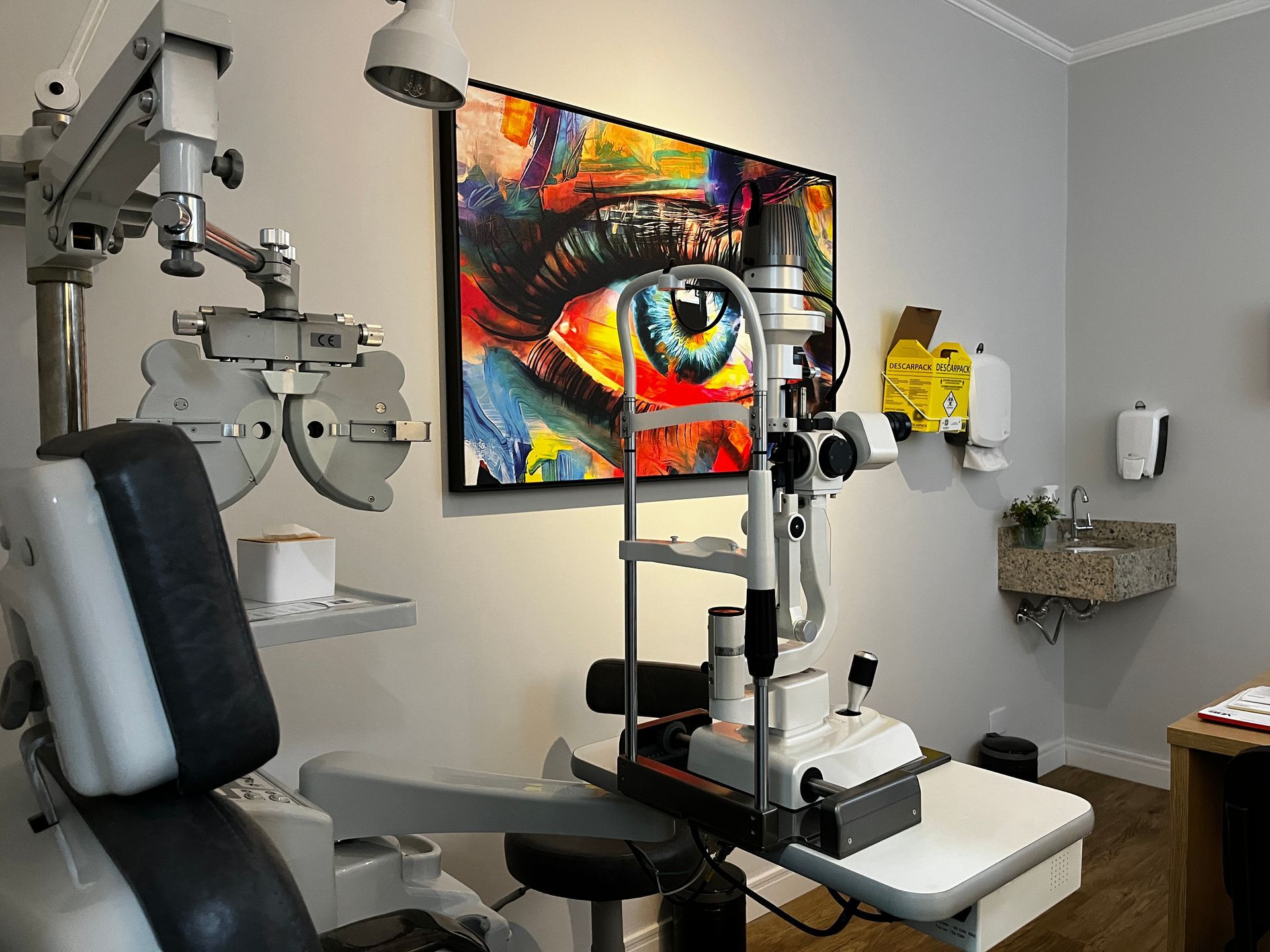 Sala de exame de optometria com equipamentos, obras de arte coloridas com o tema de olhos e pia.