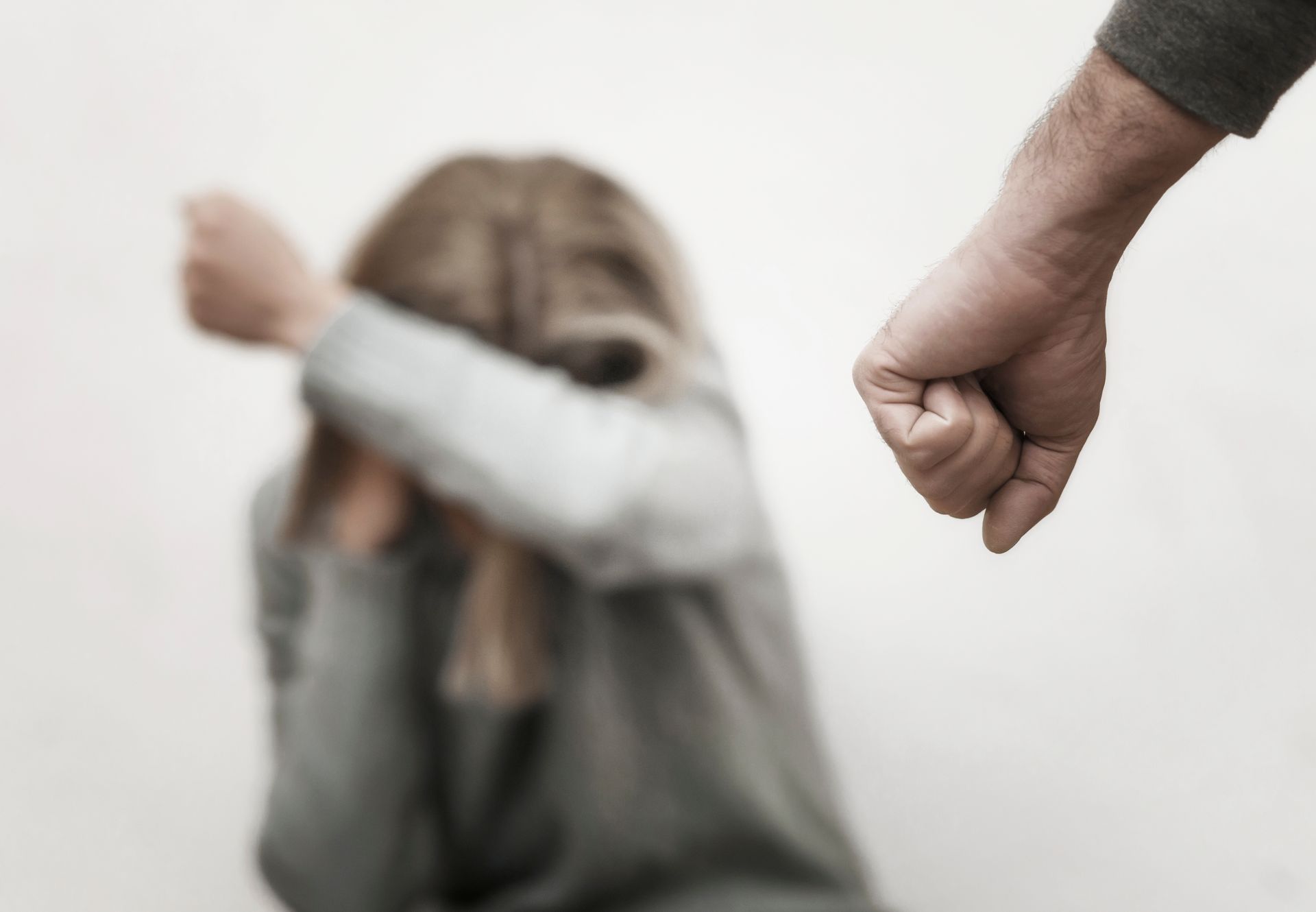 Fist raised towards person covering face with arms, appearing distressed; light background.