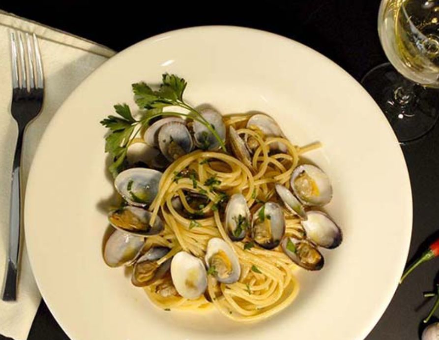 Un piatto bianco con sopra spaghetti e vongole accanto a un bicchiere di vino