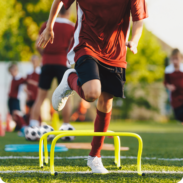 A soccer player is jumping over a yellow hurdle on a field.