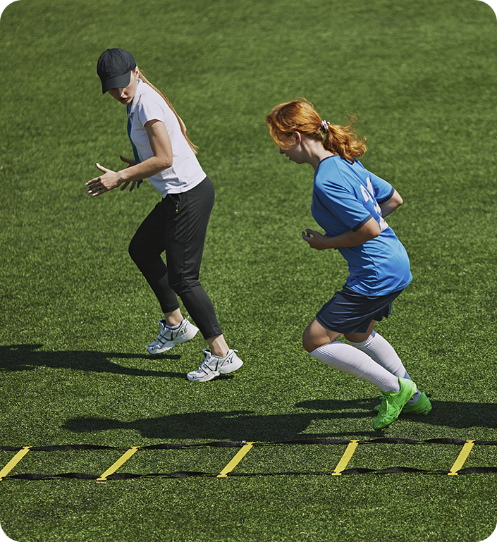Two women are jumping over a ladder on a soccer field.