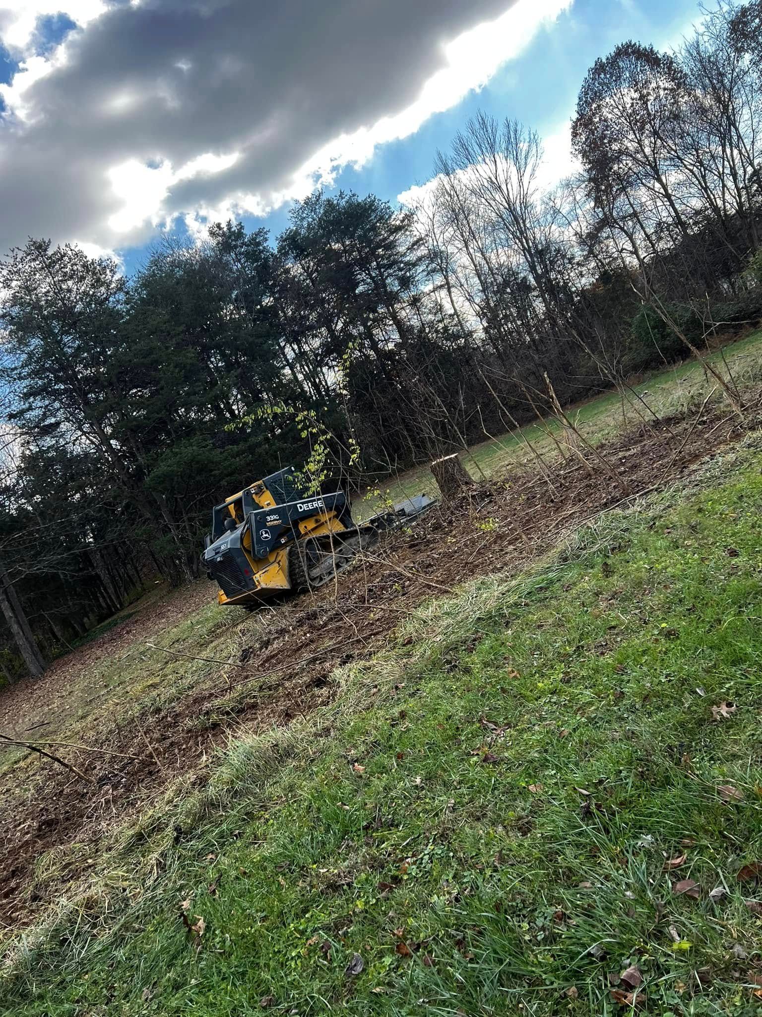 A yellow excavator clearing a grassy field, trees in the background, under a cloudy sky.