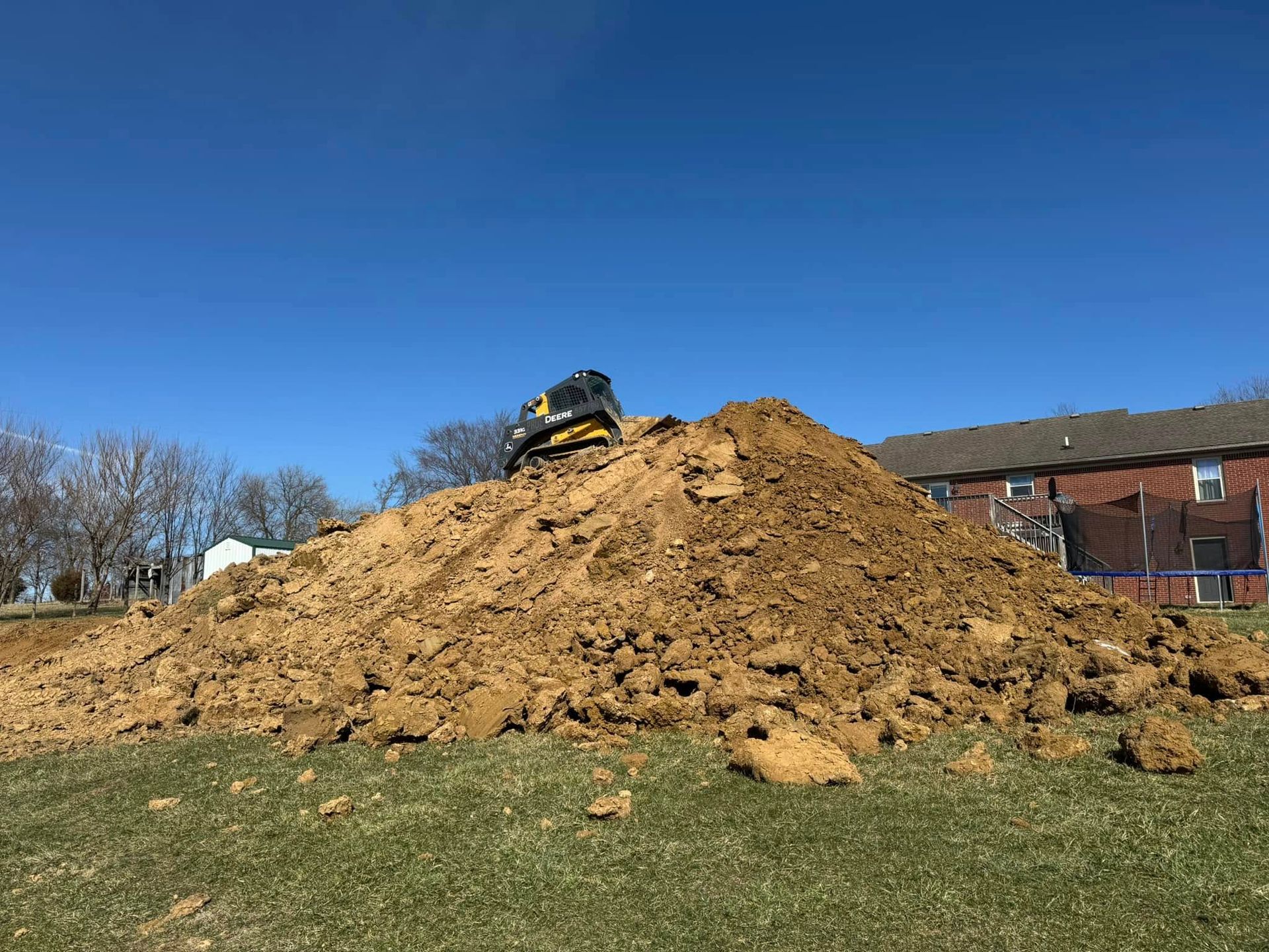 Large pile of brown dirt on a grassy field with a small construction vehicle on top, blue sky, and a building in the background.