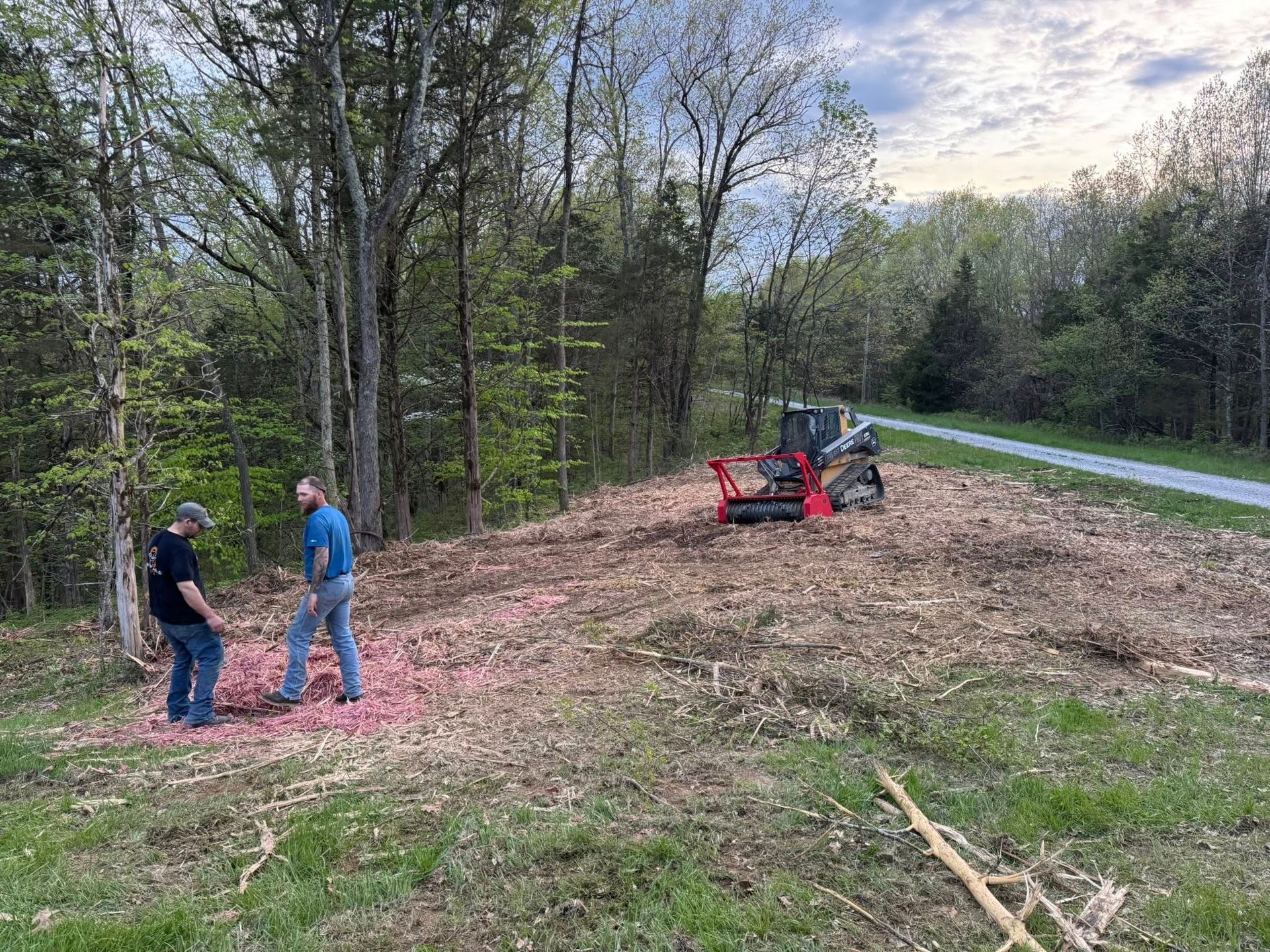 Two men stand near a mulched area; a small excavator is visible. A path and trees are in the background.
