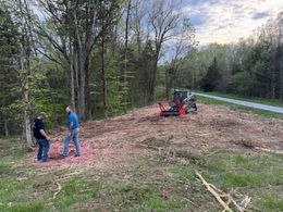 Two men stand near a mulched area; a small excavator is visible. A path and trees are in the background.