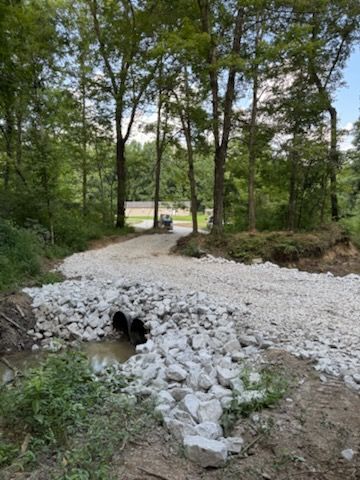 Gravel road over a stream with two pipes. Surrounded by trees, bushes and light gray rocks.