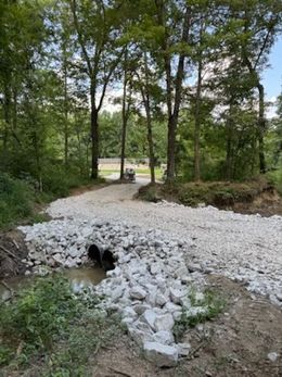 Gravel road over a stream with two pipes. Surrounded by trees, bushes and light gray rocks.