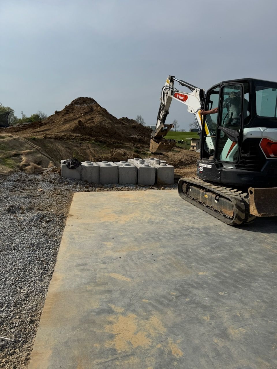 A small excavator placing concrete blocks. Pile of dirt in the background. Outdoor daytime setting.