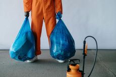 A man in an orange jumpsuit is holding two blue trash bags next to a sprayer.