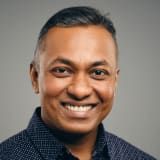 Man smiling at the camera, wearing a dark blue dotted shirt, against a gray background.