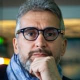 Man with graying hair and beard wearing glasses, resting chin on hand, looking at camera.