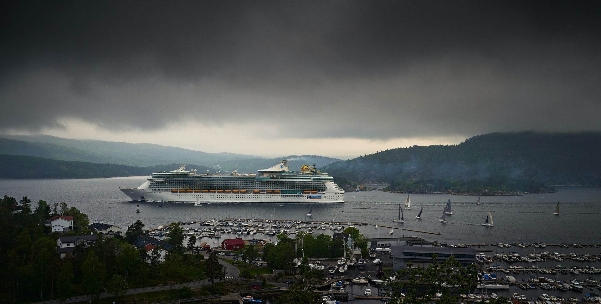 Cruise ship sails in fjord under a cloudy sky near a coastal town.